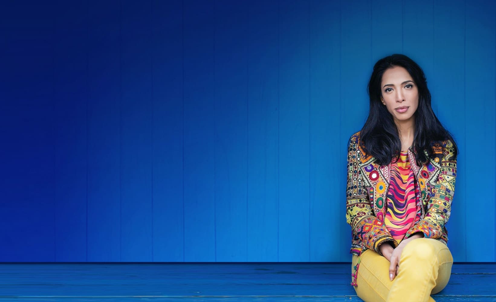 Zohre Esmaeli woman with black hair sitting in a colorful patterned jacket and yellow pants in front of a blue background