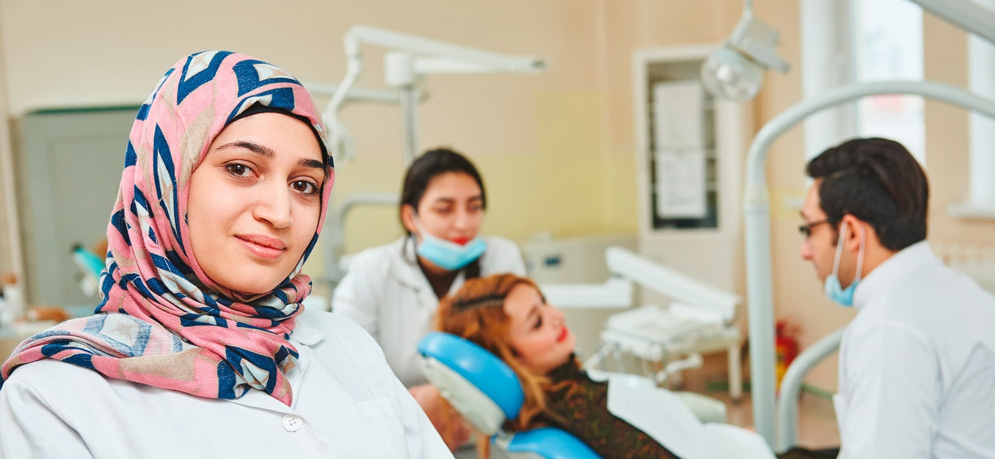 Frau mit Kopftuch im Arztkittel im Vordergrund - im Hintergrund Patientin auf Zahnarztstuhl